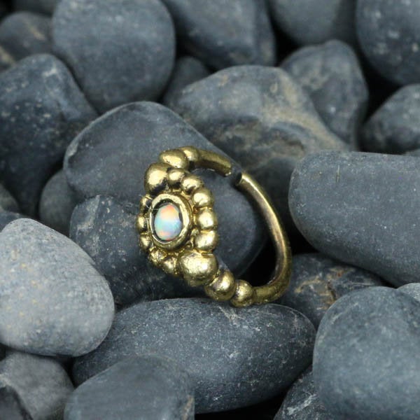 A golden nose ring featuring a central black onyx gemstone and a beaded flower design is placed on a bed of dark, smooth pebbles. The SHA Beaded Flower Nose Ring in Gold & Black Onyx by Kaya Movement showcases an ethnic-inspired design surrounding the central stone.